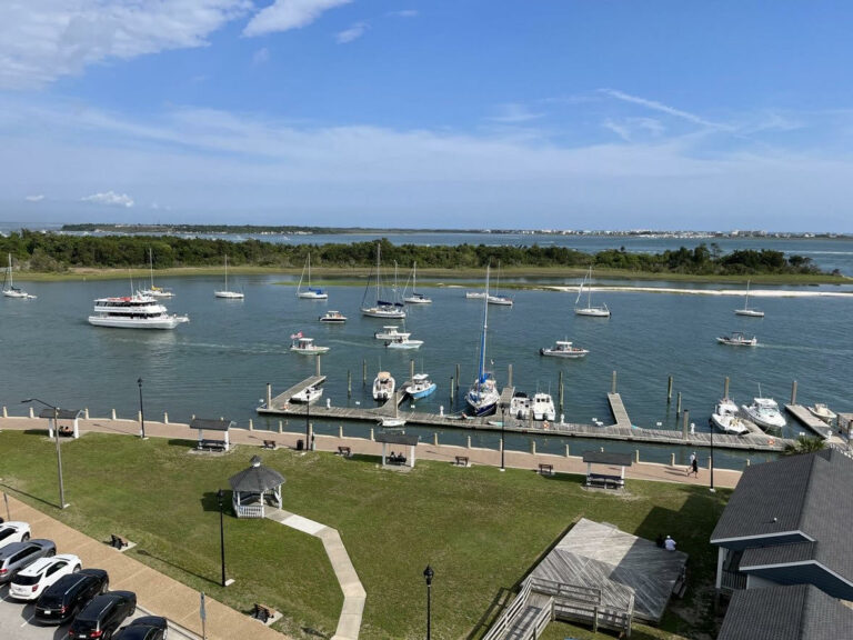 A view of a marina with several boats docked and sailing on the water, a grassy area with a gazebo, and a parking lot with cars in Morehead City, NC. The sky is clear with a few clouds.