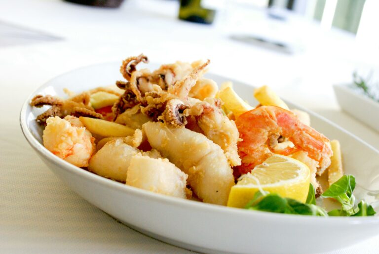 A close-up of a bowl filled with assorted fried seafood, including shrimp, calamari, and fish, garnished with lemon wedges and greens.